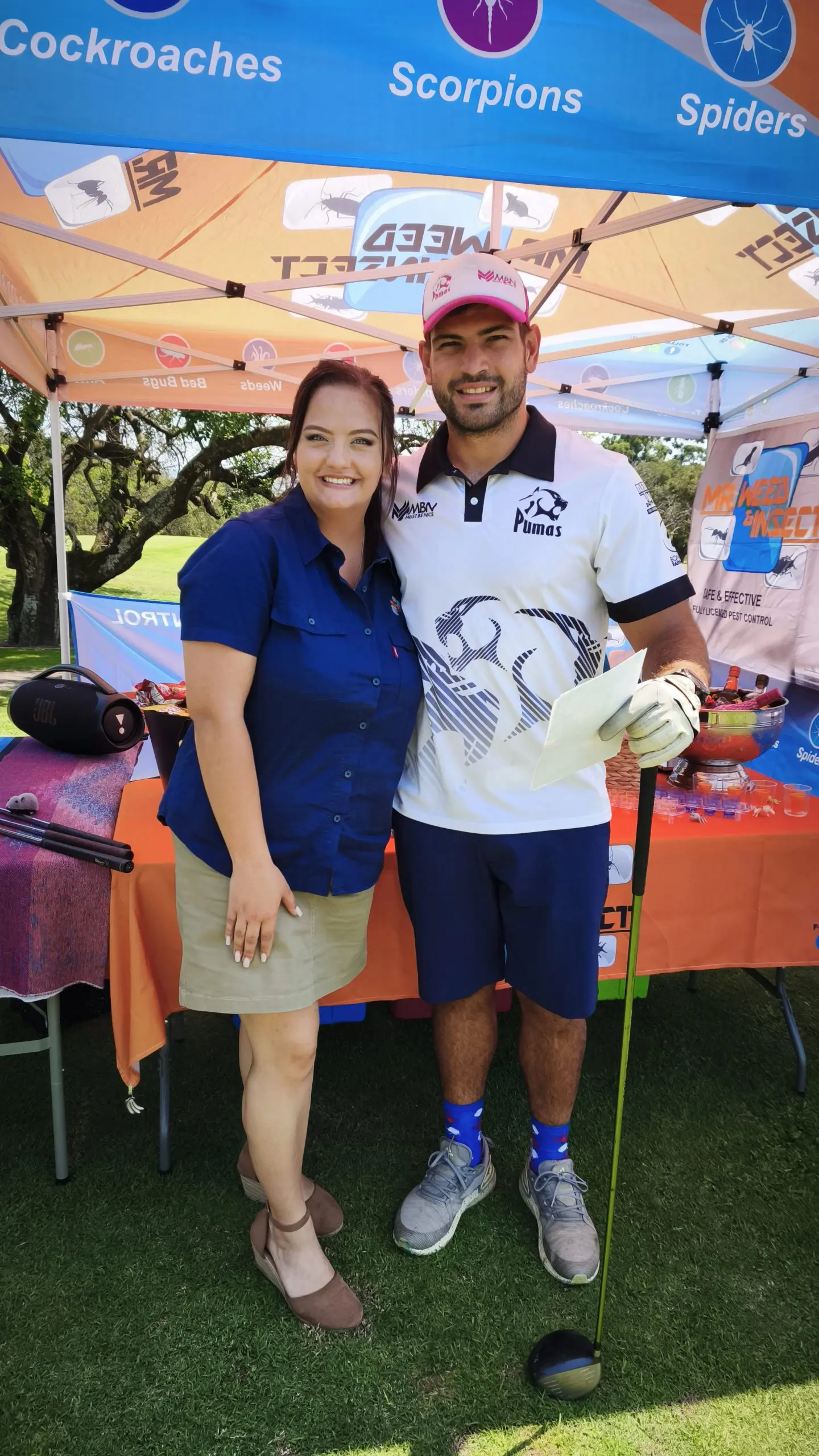 Woman in a blue shirt standing beside a man wearing a Pumas golf shirt and holding a golf club under a branded tent.