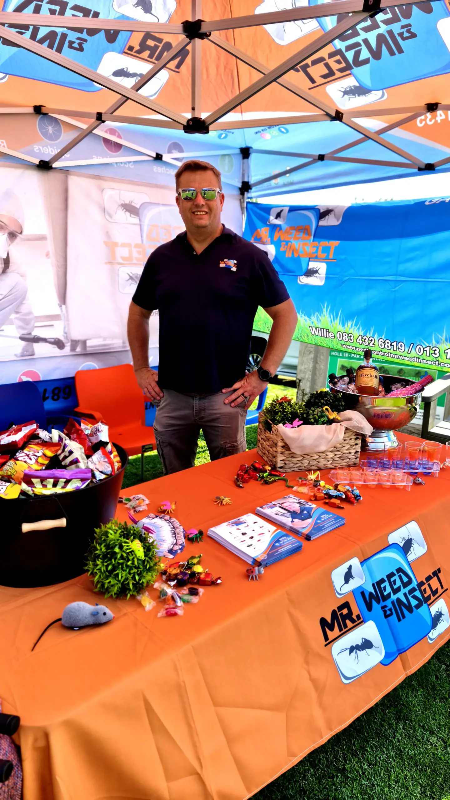 Man wearing sunglasses stands under a branded Mr. Weed & Insect canopy at an outdoor event table with snacks and brochures.