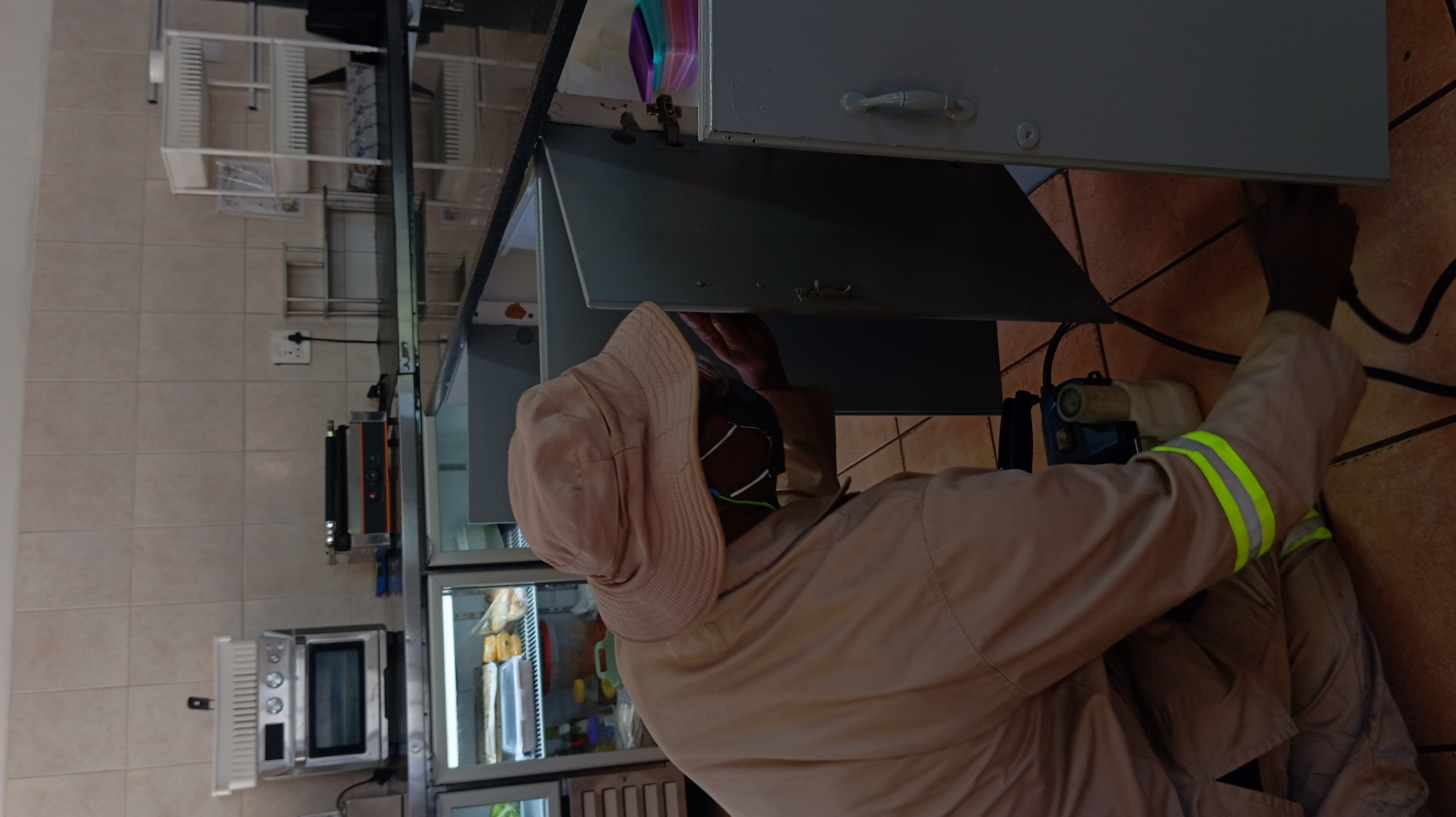 Person in a khaki uniform and bucket hat inspects the interior of a grey cabinet in a kitchen setting.