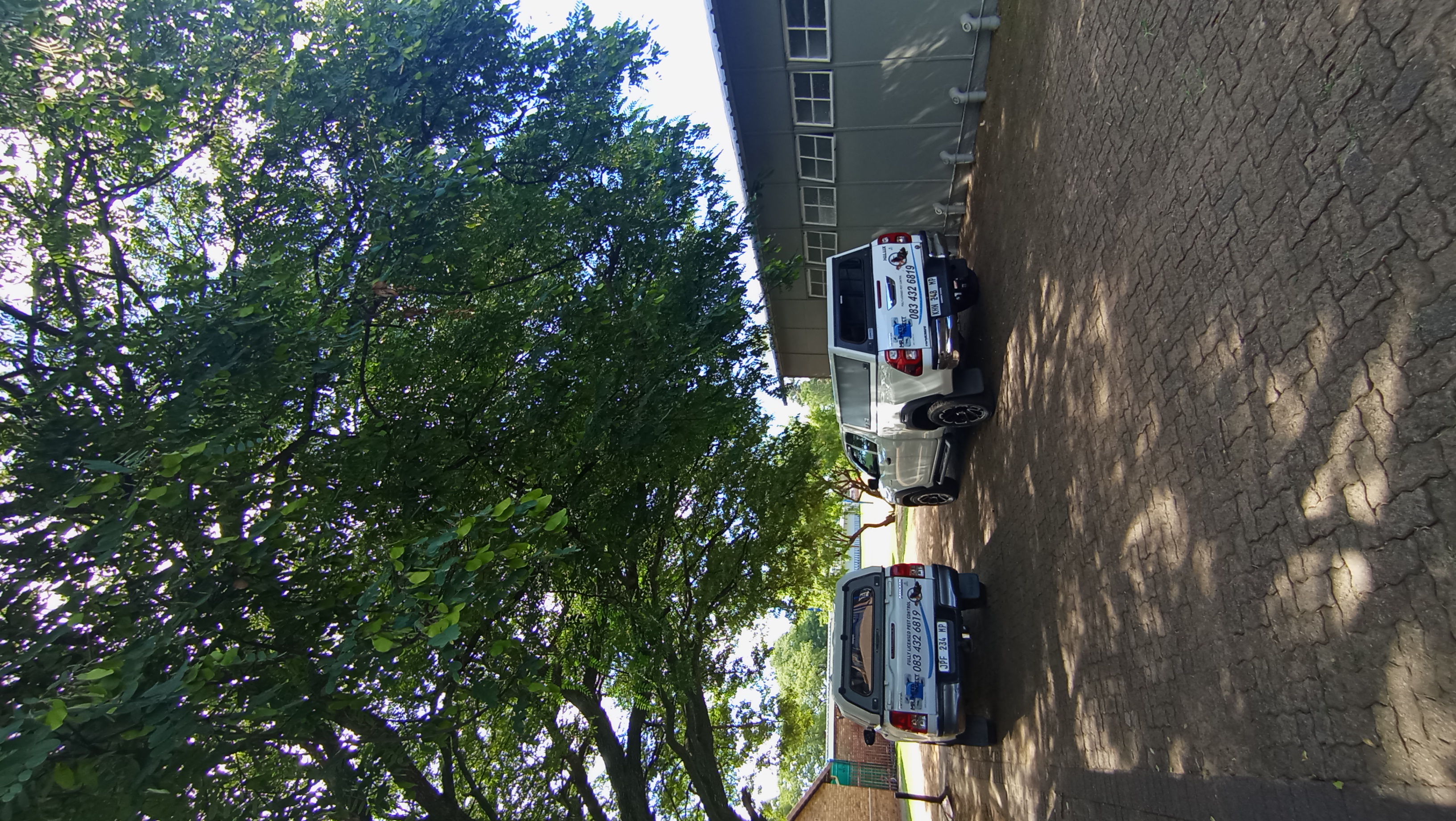 Two white pickup trucks with canopies parked in a paved lot alongside a large industrial building and leafy trees.