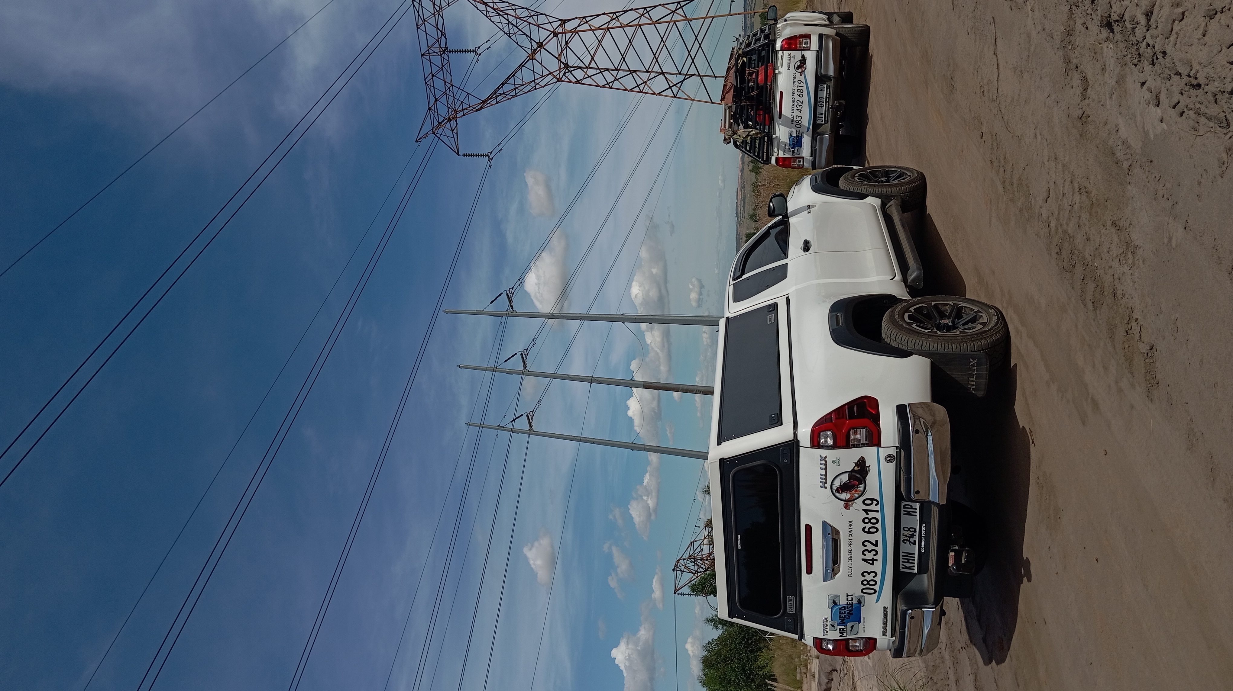 White Toyota Hilux pickup truck parked on a dirt road beneath massive steel high-voltage electrical transmission towers.