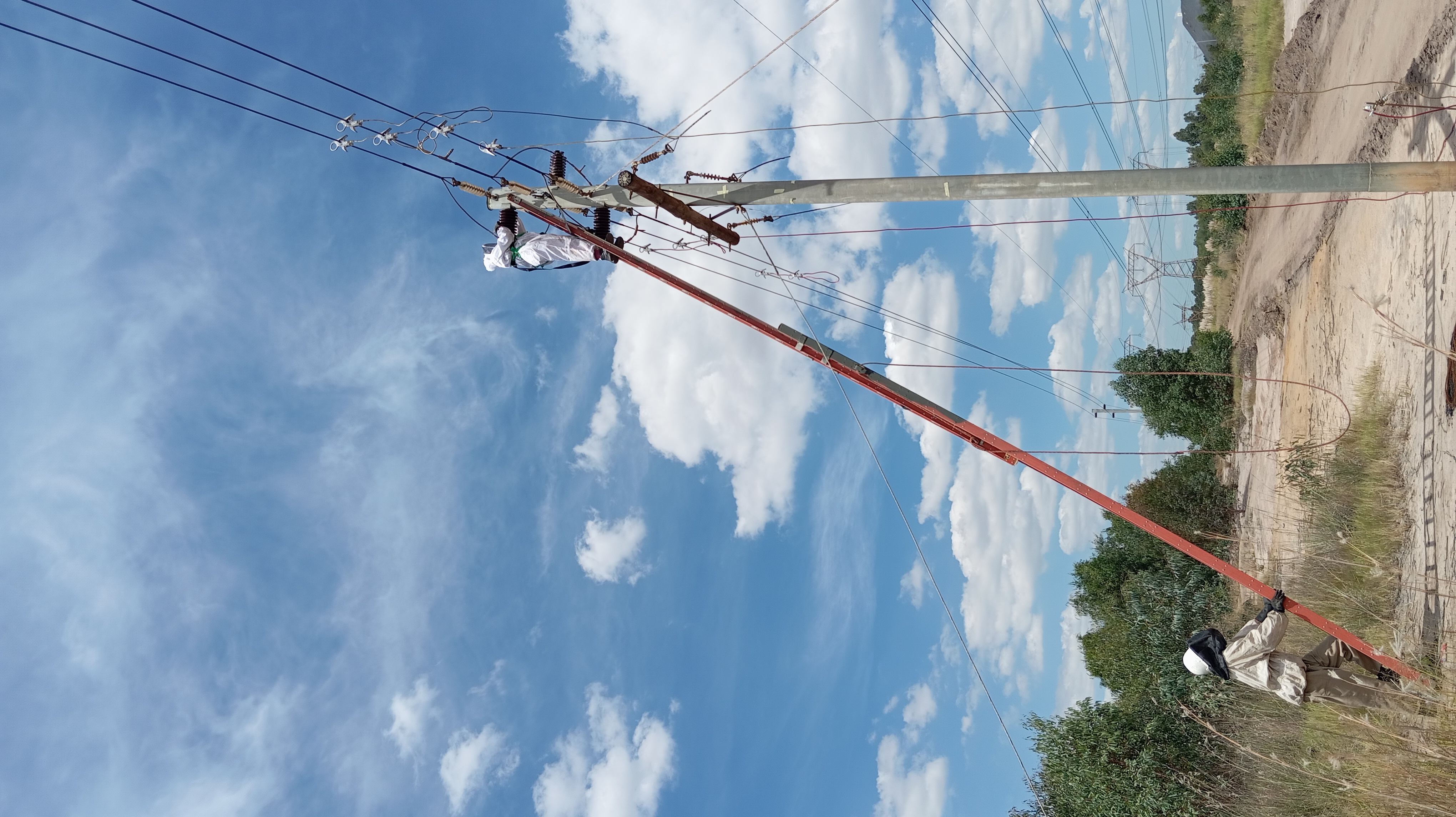 Utility worker in protective gear climbs a ladder to repair power lines on a concrete pole under a blue sky.
