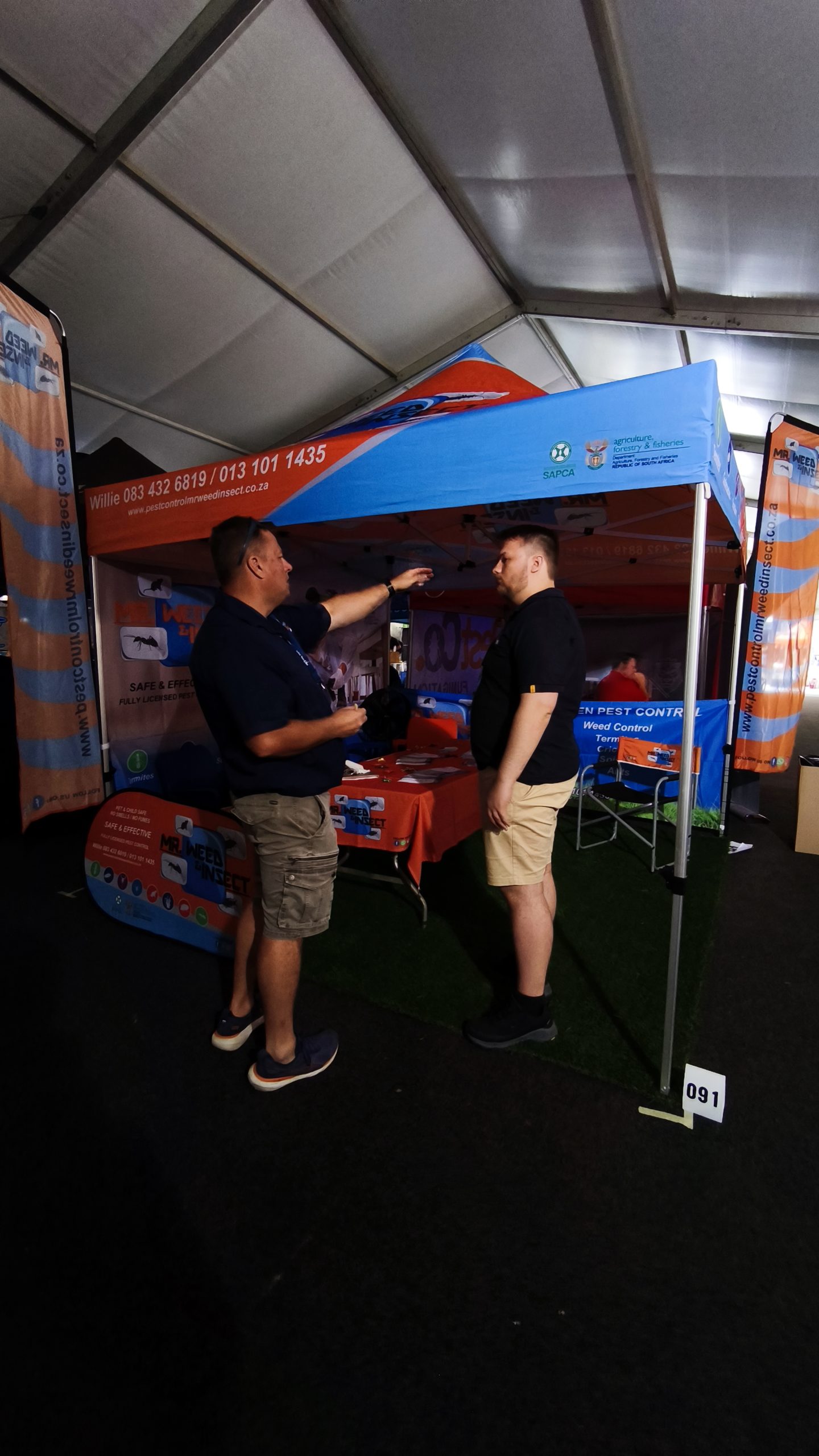 Two men stand inside a branded Mr Weed Insect exhibition tent and discuss pest control services at an event.