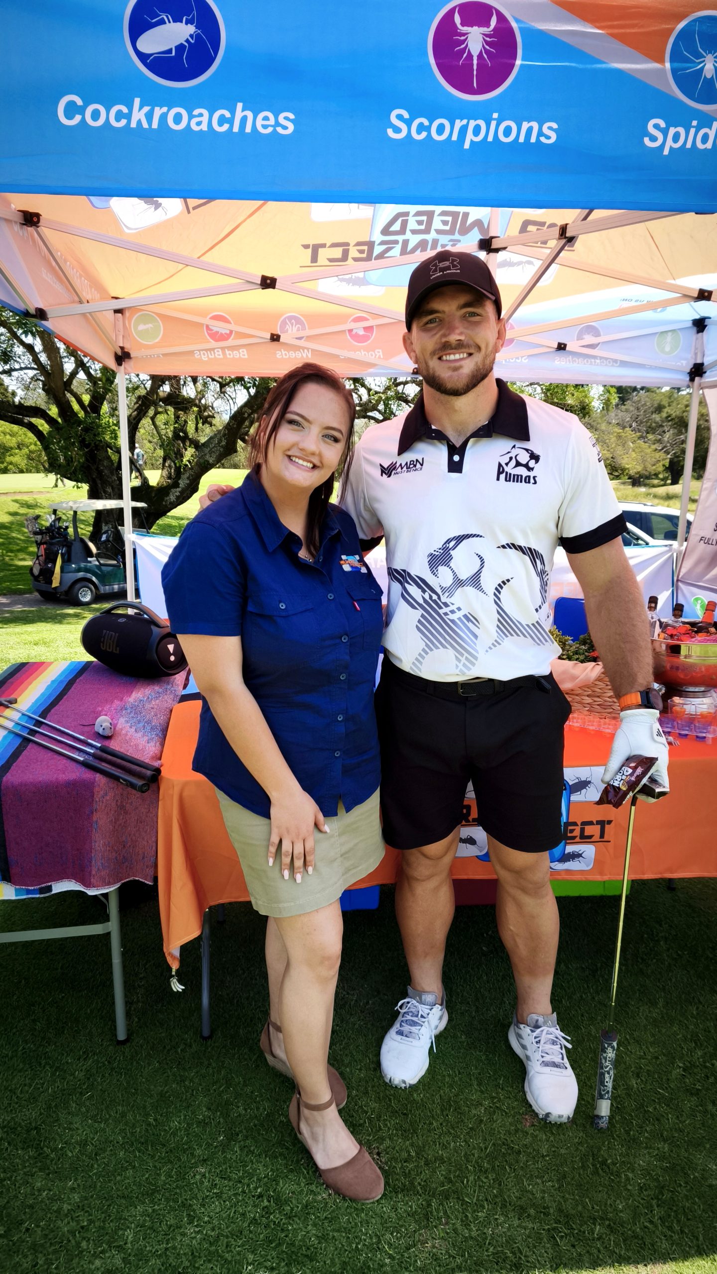 A woman and a man in a Pumas golf shirt stand smiling under a pest control tent at a golf course.
