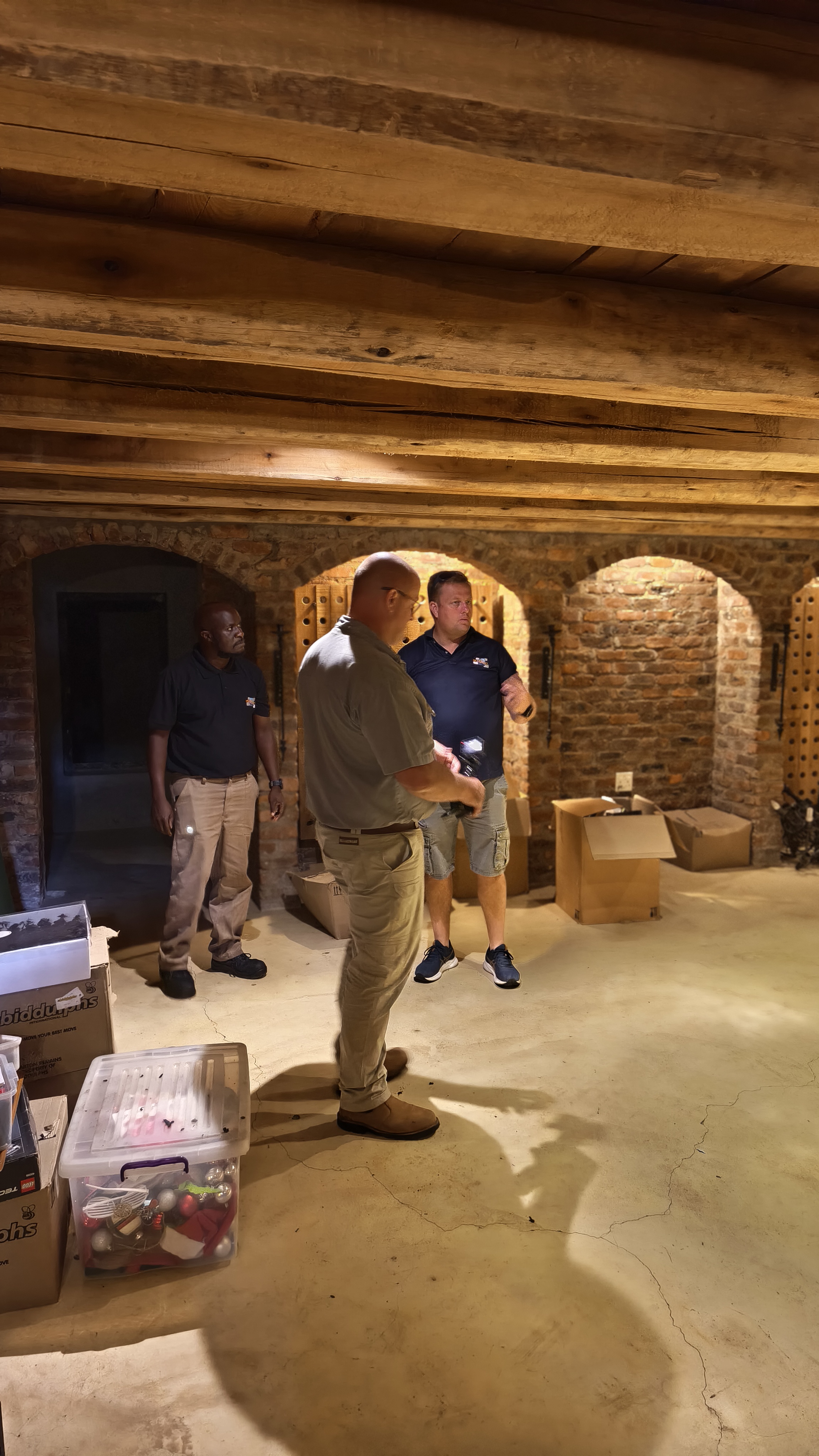 Three men stand in a rustic cellar with exposed wooden ceiling beams and arched brick walls during a storage project.