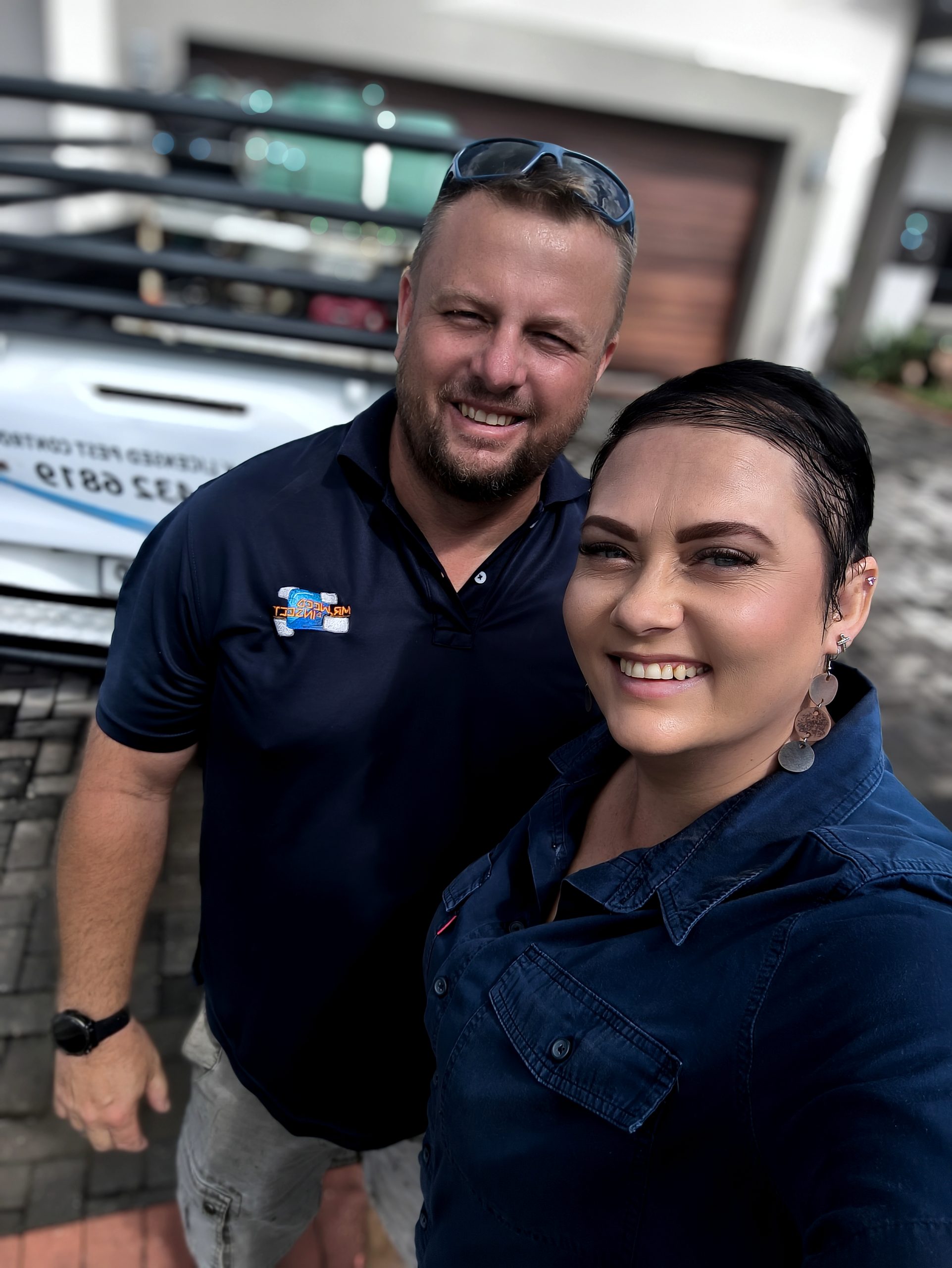 A man and a woman smiling for a selfie outdoors in front of a white work truck with a visible company logo.