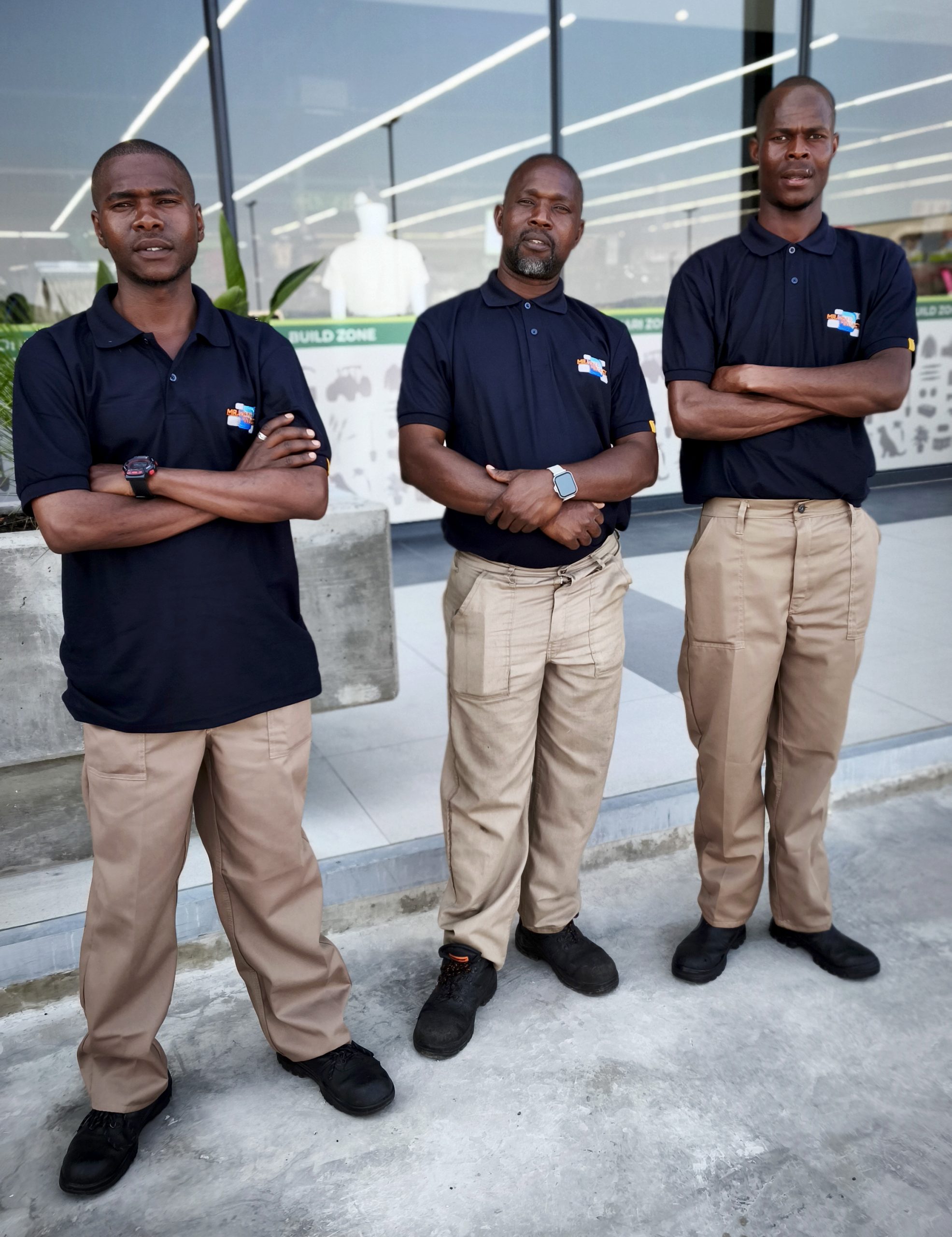 Three men in matching dark blue polo shirts and tan trousers stand with arms crossed outside a building.