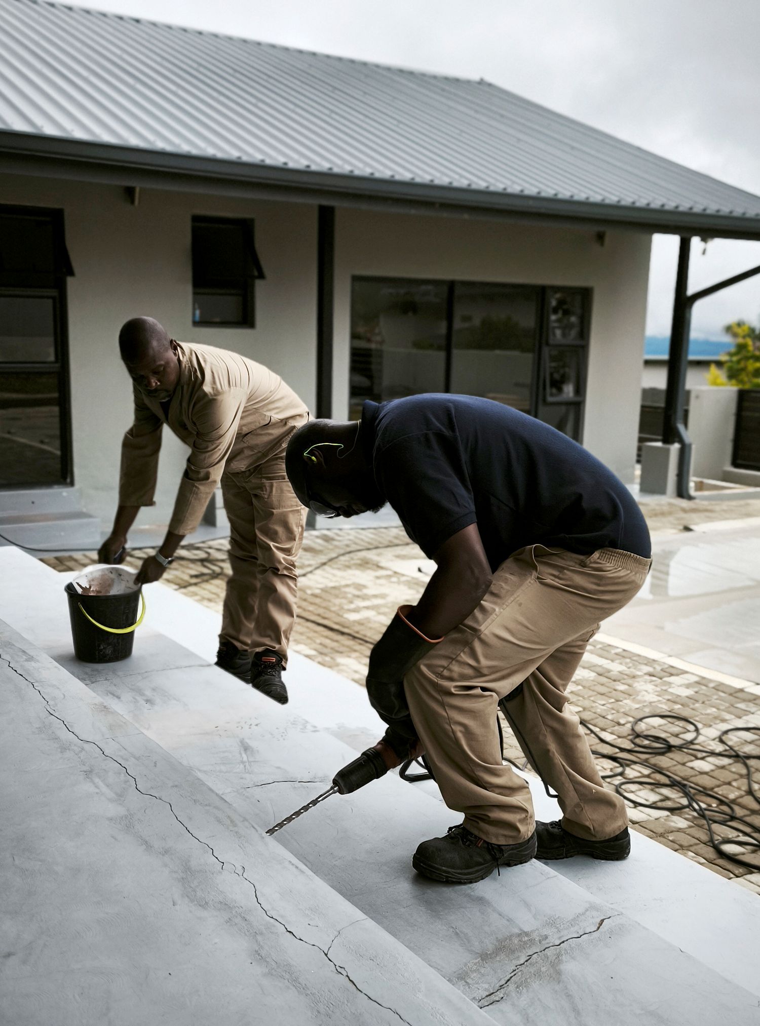 Two men in work uniforms perform maintenance on concrete steps outside a house, one drilling and the other holding a bucket.