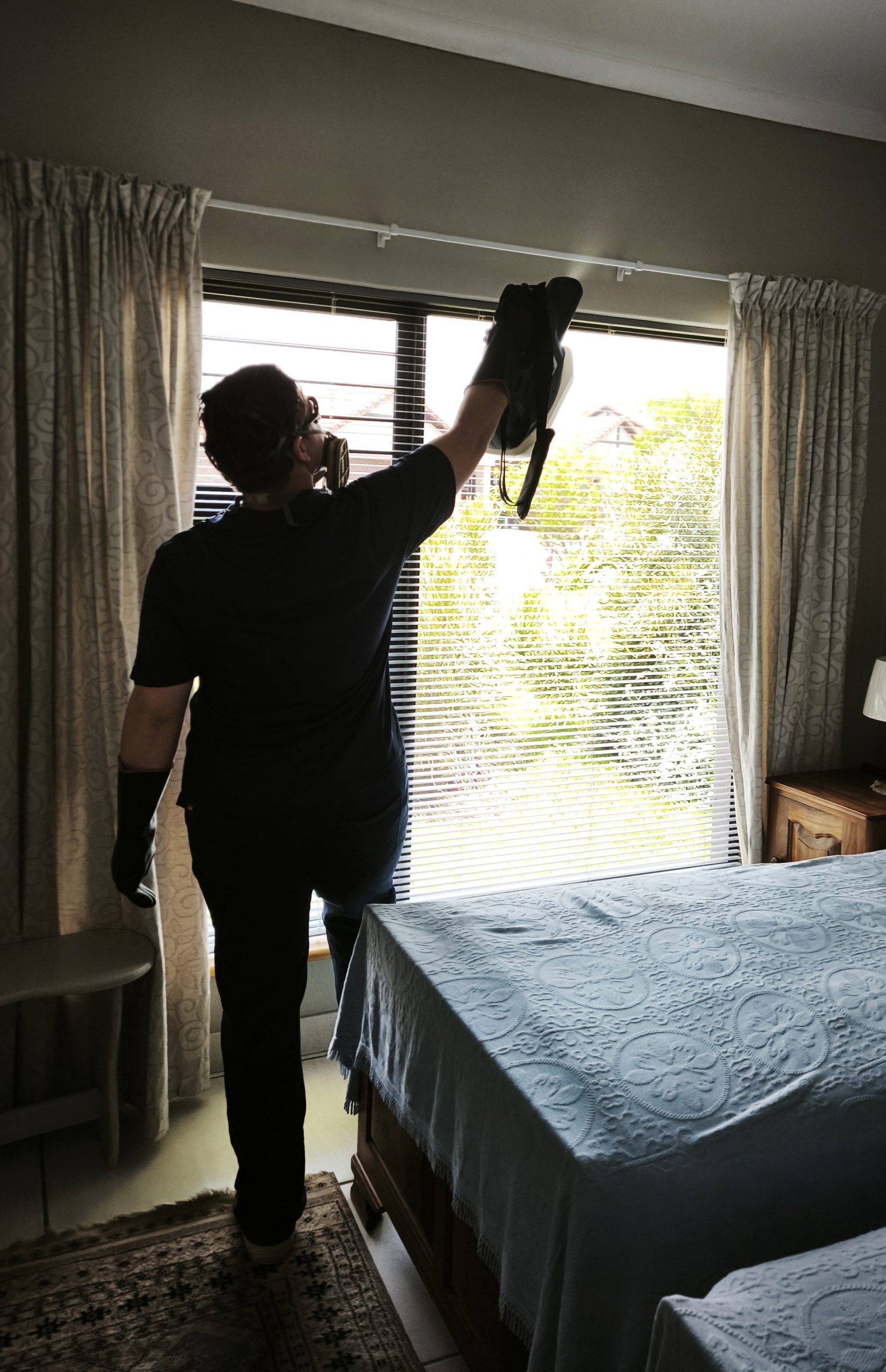 Person wearing a respirator and black gloves spraying a chemical treatment near a bedroom window.