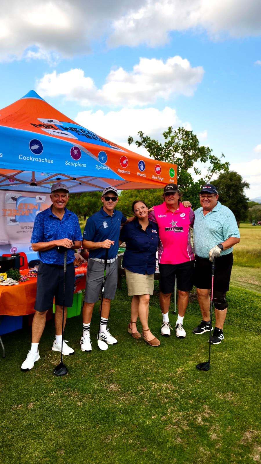 Four golfers and a woman pose in front of a blue and orange Mr. Weed and Pest canopy tent on a sunny golf course.