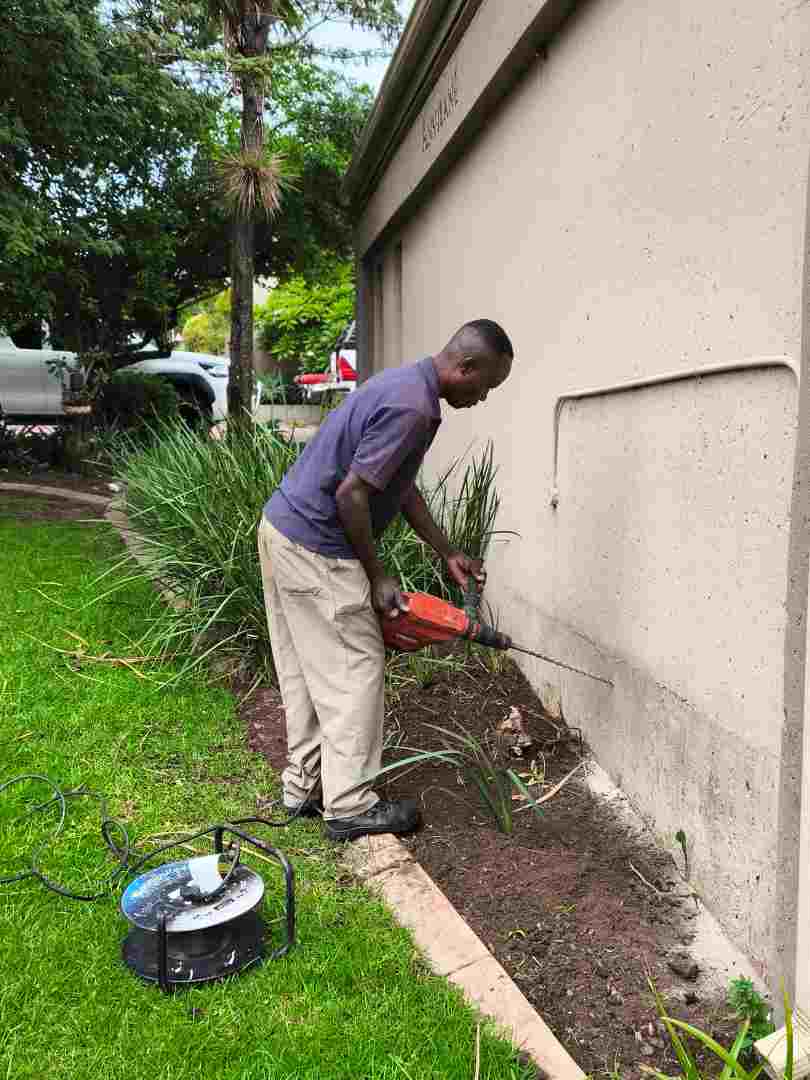 Man in a blue shirt and khaki pants drills into the concrete foundation of a beige exterior wall.
