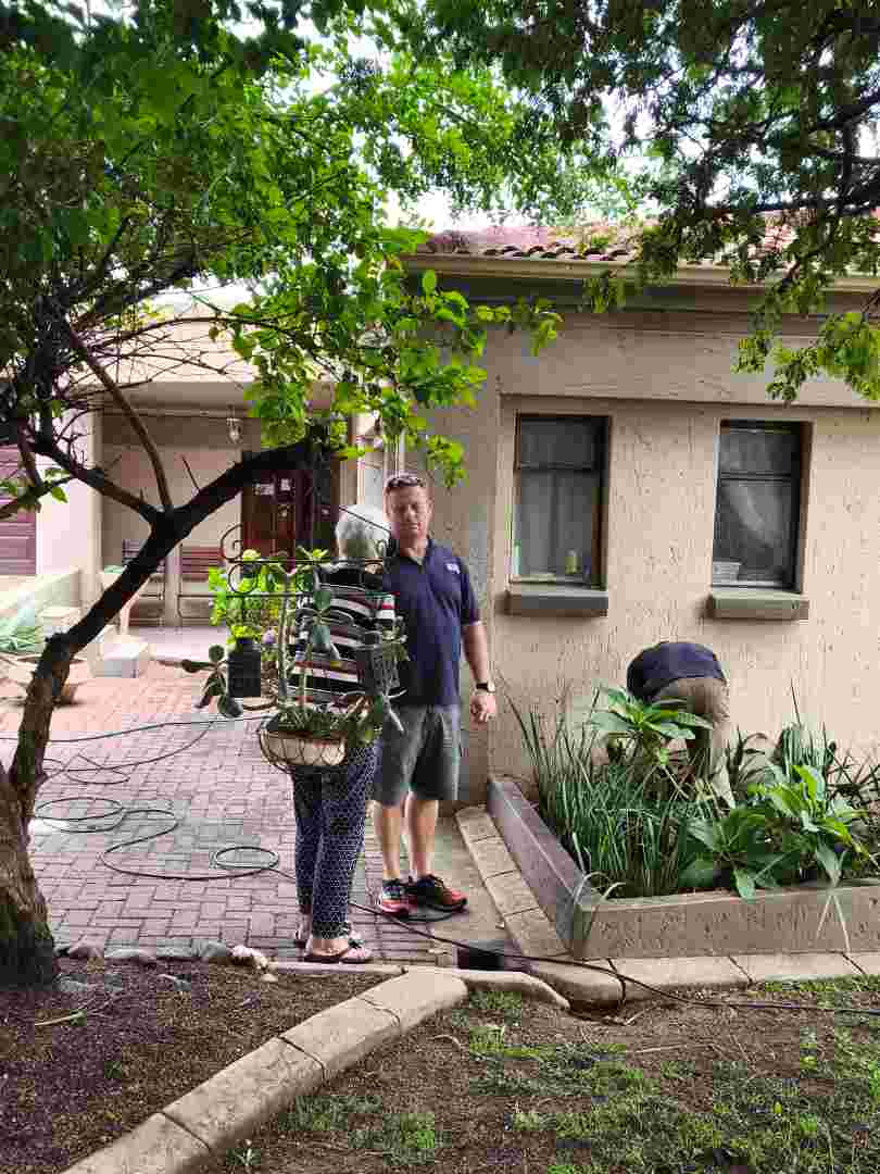 A man stands on a brick patio next to a woman while a second man works in a garden bed near a beige house.