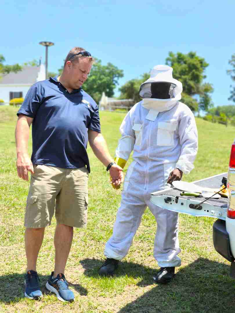 Man in a navy polo shirt talking to a person wearing a white beekeeping protective suit in a sunny outdoor field.