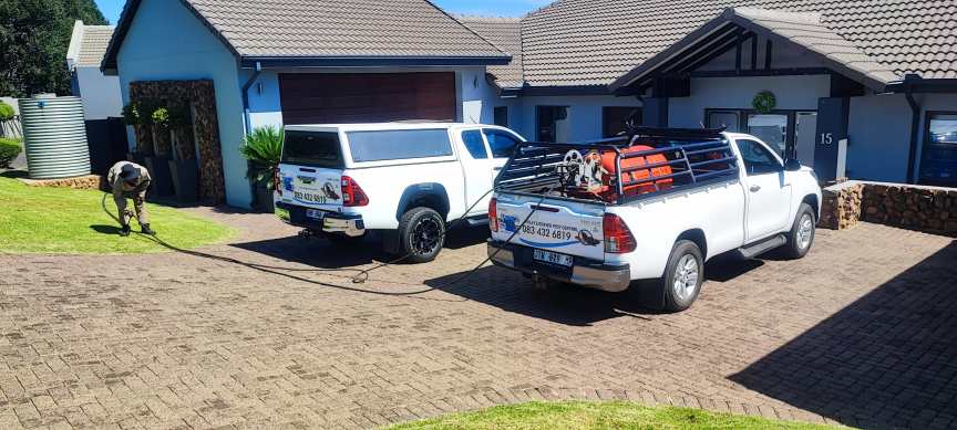 Worker in a tan uniform sprays a lawn with a hose connected to a white pickup truck parked in a residential driveway.